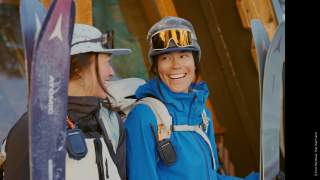 Zwei lächelnde Frauen in Skibekleidung und Helmen stehen mit aufrechtem Ski vor einer Holzhütte.