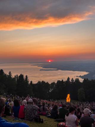 Viele Menschen sitzen auf einer Wiese des Pfänders vor einem großen Feuer. Währenddessen betrachten sie hinter einer Baumkante den Sonnenuntergang über dem Bodensee.
