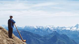 Ein Mann in traditioneller Kleidung steht auf einem Hang und spielt auf einem Alphorn mit Blick auf schneebedeckte Berge und ein Tal.