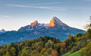 Markante Doppelspitze der Watzmanner Berglandschaft mit herbstlich buntem Wald im Vordergrund und schneebedeckten Gipfeln, die im warmen Licht der untergehenden Sonne leuchten.