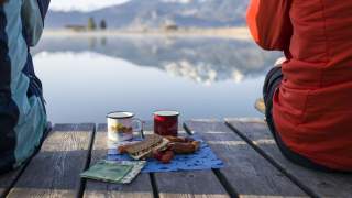 Zwei Personen sitzen auf einem Holzsteg am See, zwischen ihnen liegt ein kleiner Picknickaufbau mit Sandwich, Brezel, Wurst, Tomaten und zwei Bechern. Im Hintergrund spiegeln sich Berge im Wasser.