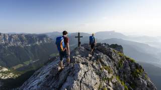 Zwei Wanderer mit Rucksäcken gehen auf einem felsigen Berggipfel auf ein Gipfelkreuz zu, umgeben von einem weiten Bergpanorama.