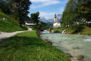 Über einen kleinen Fluss führt eine Holzbrücke, dahinter eine Kirche, im Hintergrund hohe Berge.