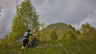 Ein Radfahrer mit Helm und Rucksack schiebt sein Mountainbike einen grasbewachsenen Hang hinauf, im Hintergrund ein Hügel mit einem Gipfelkreuz unter bewölktem Himmel.