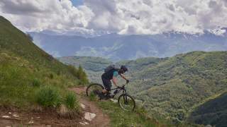 Ein Mountainbiker fährt auf einem schmalen, kurvigen Pfad an einem grünen Berghang mit Blick auf bewaldete Hügel und schneebedeckte Berge unter wolkigem Himmel.
