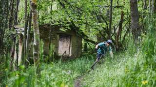 Ein Mountainbiker fährt auf einem schmalen Waldpfad vorbei an einer kleinen, verfallenen Steinhütte im grünen Frühjahrwald.