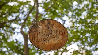Holzschild mit der Aufschrift „DAHU“ hängt an einer Kette vor einem verschwommenen grünen Laubhintergrund.