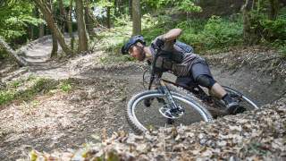 Ein Mann mit Helm und Schutzkleidung fährt in einem Wald auf einem Mountainbike eine kurvige, mit Laub bedeckte Dirt-Trail-Strecke. Er lehnt sich beim Fahren stark in die Kurve.