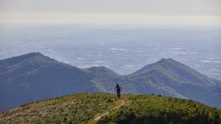 Eine Person fährt mit Mountainbike auf einem schmalen Pfad über einen grasbewachsenen Hügel mit bergiger Landschaft im Hintergrund.
