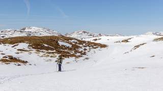 Eine einzelne Person beim Skitourengehen auf verschneitem Gelände mit vereinzelten Schneeflecken und kahlen Hügeln, im Hintergrund schneebedeckte Berge unter blauem Himmel.