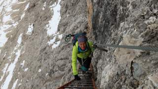 Person mit Kletterausrüstung steigt eine Metallleiter an einer steilen, felsigen und teilweise verschneiten Bergwand hinauf.