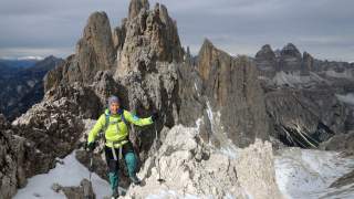 Eine Person mit Wanderkleidung und Stöcken steigt auf einem schneebedeckten, felsigen Gebirgspfad, im Hintergrund steile, zerklüftete Berge unter bewölktem Himmel.