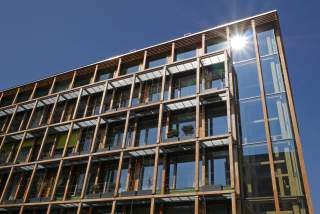 Fassade eines modernen Gebäudes mit großen Glasflächen und Holzrahmen, die Sonne spiegelt sich in einer Fensterscheibe. Mehrere kleine Pflanzen sind auf den Balkonen zu sehen.