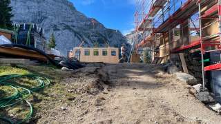 Bauarbeiter montieren im Gebirge mit Hilfe eines Krans eine Holzfassadenwand an einem Gerüst neben einem Baugelände. Im Hintergrund sind steile Felswände unter blauem Himmel zu sehen.