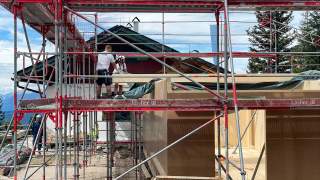 Bauarbeiter mit Schutzhelmen stehen auf einem Gerüst und arbeiten an einem Holzbau vor einer traditionellen Berghütte mit grünem Dachfirst, umgeben von Bergen und Bäumen.