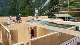Mehrere Bauarbeiter mit Schutzhelmen montieren Holzwände und Balken auf einer Baustelle in bergiger Waldlandschaft.