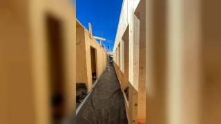 Baustelle mit aufgestellten Holzrahmen für Wände und offenem Gang dazwischen, im Hintergrund steht eine Stehleiter unter blauem Himmel.