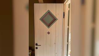 helle Holztür mit vertikalen Lamellen, darauf zwei ineinandergelegte quadratische Fensterrahmen in Grün und Rot sowie ein kleiner roter quadratischer Akzent darunter. Schwarzer Türgriff und Schlüsselloch befinden sich links unten.