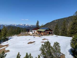 Berghütte mit Solarpanelen auf dem Dach in einer schneebedeckten Alpenlandschaft, umgeben von Tannen und Bergen unter klarem blauem Himmel.