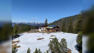 Große weiße Säcke mit rotem Band, gefüllt mit verschiedenen Materialien, stehen im Freien vor schneebedeckten Bergen und Nadelbäumen, daneben eine Person in schwarzer Kleidung.