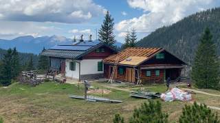 Berghütte mit zwei Gebäudeteilen, das Haupthaus mit Solarpanelen auf dem Dach und das Nebengebäude mit Dachrückbau, umgeben von Bergen und Wäldern. Zwei Personen arbeiten auf dem Gelände, Material und Leitern sind sichtbar.
