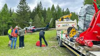 Menschen stehen auf einer Wiese neben einem geparkten Helikopter und einem mit Baumaterialien und Maschinen beladenen Lastwagen, umgeben von Nadelbäumen unter bewölktem Himmel.