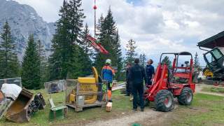 Mehrere Arbeiter an einer Baustelle in den Bergen beobachten ein mit einem Helikopter eingeflogenes Teil eines Baggers. Im Hintergrund sind Tannenbäume und Berge unter einem bewölkten Himmel zu sehen.