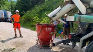 Drei Bauarbeiter stehen neben einer Betonmischmaschine und einem roten Betonbehälter auf einem Waldweg in den Bergen.