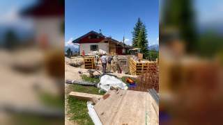 Bauarbeiter mit Schutzhelmen arbeiten an einer Baustelle vor einer Berghütte, umgeben von Baustoffen und Werkzeugen. Im Hintergrund sind Berge und ein klarer blauer Himmel zu sehen.
