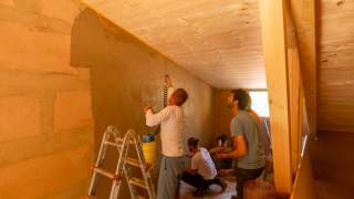 Vier Männer verputzen eine Innenwand in einem Raum mit Holzbalken und Decke mit Lehm, im Vordergrund steht eine Metallleiter.