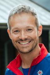 Henrik Vogel von der Bergwacht München Ein Mann von der Bergwacht München schaut in die Kamera und lächelt. Er steht vor der Diensthütte und trägt seine Bergwacht-Kleidung in den Farben rot und blau.