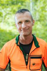 Bernhard Reissner ist Förster im Mangfallgebirge in den Bayrischen Voralpen Ein Mann trägt seine orange-grünen Dienstkleidung mit dem Logo der Bayerischen Staatsforsten auf der Brusttasche. Er steht im Wald, umgeben von Bäumen. Er blickt lächelnd in die Kamera.