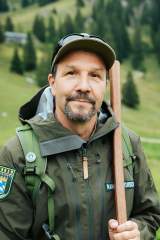 dav_bergader_reels_ranger.jpg Der Ranger Alex Römer steht in seiner grünen Dienstkleidung auf einer Bergwiese. In der Hand hält er einen Holzstab.