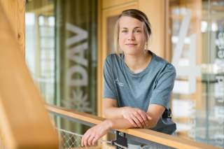 Frau lehnt an einem Geländer aus Holz und Metall in einem modernen, hellen Innenraum und blickt freundlich in die Kamera. Sie trägt ein graues T-Shirt mit einem Logo auf der Brust.