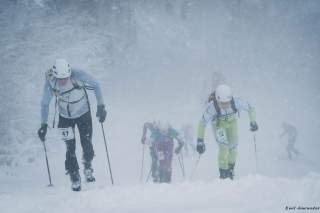 Mehrere Skifahrer mit Helmen und Ausrüstung kämpfen sich bei starkem Schneefall einen verschneiten Hang hinauf.