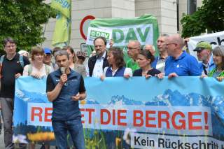 Eine Person im Vordergrund spricht ins Mikrofon, im Hintergrund sind mehrere Personen hinter einem Banner mit der Aufschrift 
