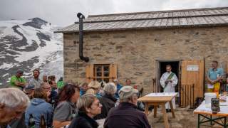 Gäste sitzen vor der Alten Prager Hütte und hören dem Pfarrer zu.