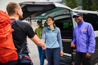 Drei Bergsportler*innen vor einem Auto mit offener Heckklappe, zwei begrüßen sich.