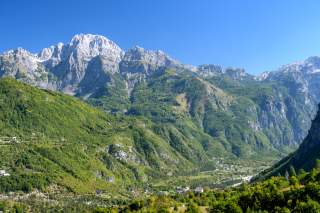 Blick auf ein grünes, bewaldetes Tal mit verstreuten Häusern und schroffen, teils schneebedeckten Bergen unter klarem blauem Himmel.
