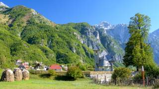 Berglandschaft mit grünen Hängen, einer kleinen Kirche mit spitzem Dach im Vordergrund und vereinzelten Häusern, darüber blauer Himmel. Rechts steht ein großer Baum neben einem Zaun.