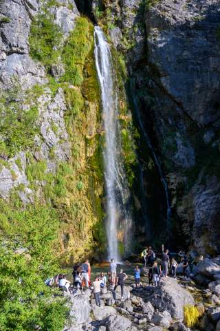 Ein Wasserfall stürzt über eine bewachsene Felswand in ein Becken, vor dem mehrere Menschen auf Felsen stehen und das Wasser beobachten.