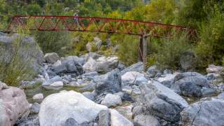 Ein Mensch läuft über eine schmale rote Metallbrücke, die über einen felsigen Bach in einer grünen, bewaldeten Landschaft führt.