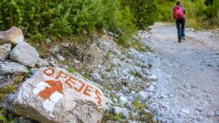 Wanderweg mit Stein, auf dem „OPEJES“ und ein roter Pfeil nach vorne gemalt sind, im Vordergrund, im Hintergrund eine Person mit rotem Rucksack, die den Weg entlanggeht.