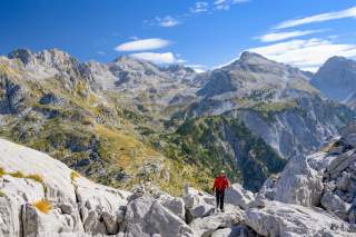 Wanderer in roter Jacke klettert auf felsigem Bergpfad in einer alpinen Gebirgslandschaft unter blauem Himmel mit vereinzelten Wolken.