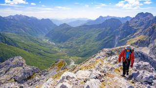 Wanderer mit roter Jacke steigt auf felsigem Bergpfad, umgeben von grünen Tälern und hohen Bergen unter blauem Himmel.