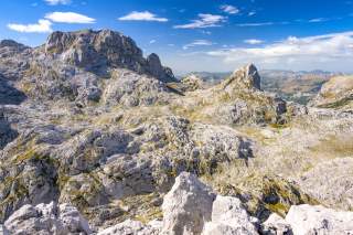 Felsige, karge Berglandschaft unter blauem Himmel mit wenigen Wolken, im Vordergrund grobe Felsen. Im Hintergrund sind Berge mit schroffen Gipfeln und leicht bewachsenen Flächen zu sehen.