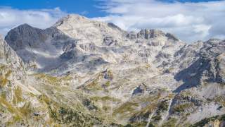 Felsige Berglandschaft mit steilen Abhängen unter einem teils bewölkten blauen Himmel. Vereinzelt sind grüne Vegetationsflächen im unteren Bereich zu sehen.