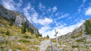 Gebirgshang mit Felsen, vereinzelten Tannen und einem hölzernen Wegkreuz unter blauem Himmel mit vereinzelten Wolken.