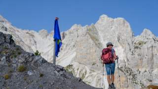 Wanderin mit rotem Rucksack und Wanderstöcken steht auf felsigem Bergpfad mit Blick auf steile, helle Felswände, daneben liegt ein Hund auf dem Boden.