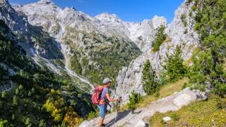 Wanderer mit rotem Rucksack und Trekkingstöcken auf einem Bergpfad, umgeben von steinigen Hängen, grünen Bäumen und hohen Bergen unter blauem Himmel.